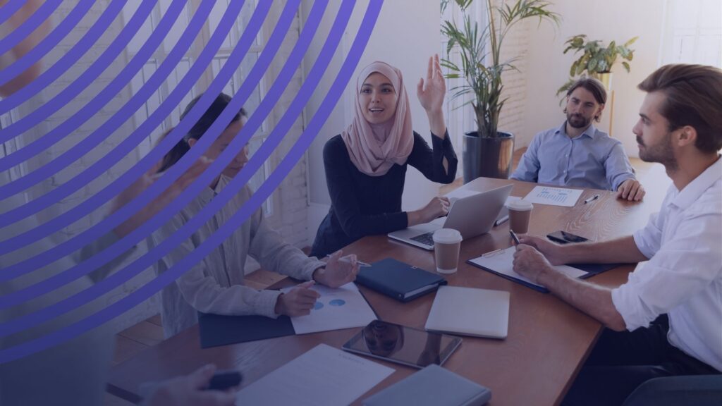 اجتماع عمل لفريق متنوع يناقش خطط التدريب والتطوير داخل الشركة، حيث تظهر سيدة محجبة ترفع يدها للمشاركة، وأمامهم أجهزة محمولة وتقارير بيانية.